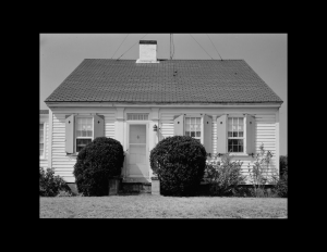 Cape cod style house with centralized chimney, double-hung windows with shutters, and shingle siding.