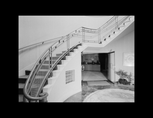 Art moderne building interior showcasing an oval first floor, with a wrap-around staircase along with rounded railings and decorative panels.