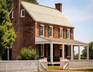 All brick farmhouse style home with steep triangular roof, window mouldings with shutters, and a covered porch with columns.