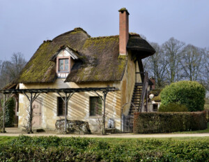 French country style cottage with stone exterior, mossy steep roof with centered gable dormer, one chimney, and outside staircase to second story.