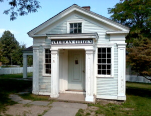 Small greek revival style building with shingle siding featuring a covered porch entrance and window mouldings.