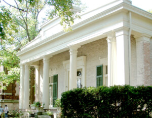 All brick greek revival style building with extensive exterior cornice mouldings, pillars, door mouldings, and windows with shutters.