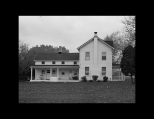 Exterior of farmhouse with side covered ground level porch, two chimneys, slanted triangular roof, and shingle siding.