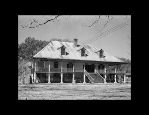 French colonial style house with steep roof, gable dormers, wrap-around balcony, covered porch underneath balcony, and windows with shutters.