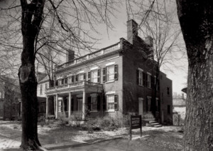 Exterior of Martin House featuring windows with shutters, door casing with column detail, covered porch with columns, and balustrades on roof.