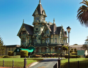 A large victorian estate sits atop a manicured landscape. The mansion features vibrant green exterior trim work with detailed carvings and embellishments. A tall central peak stretches into the skyline, creating a sense of grandeur.