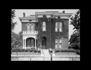 Italianate style home with hipped roof, tall window mouldings, covered walkway to entrance along with a balcony on top with balustrades.