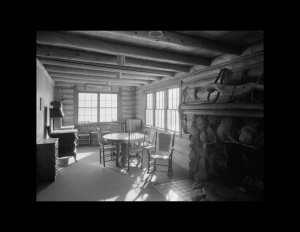 Interior of log cabin style home with many log beams going across ceiling, traditional log walls, window mouldings, and carpeted floor.