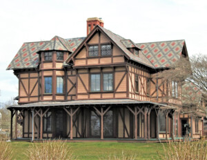 Stick style house with steep hipped roof, wrap around porch, window mouldings, brick chimney, and barn like designs.