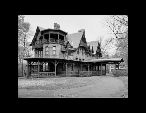 Side angle of a stick style house with hipped roof, wrap around porch, window mouldings, dormers, two brick chimneys, and balcony on top floor.