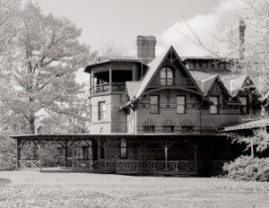 Stick style house with steep hipped roof, huge wrap around porch, window mouldings, gable dormers, two brick chimneys, and balcony on top floor.